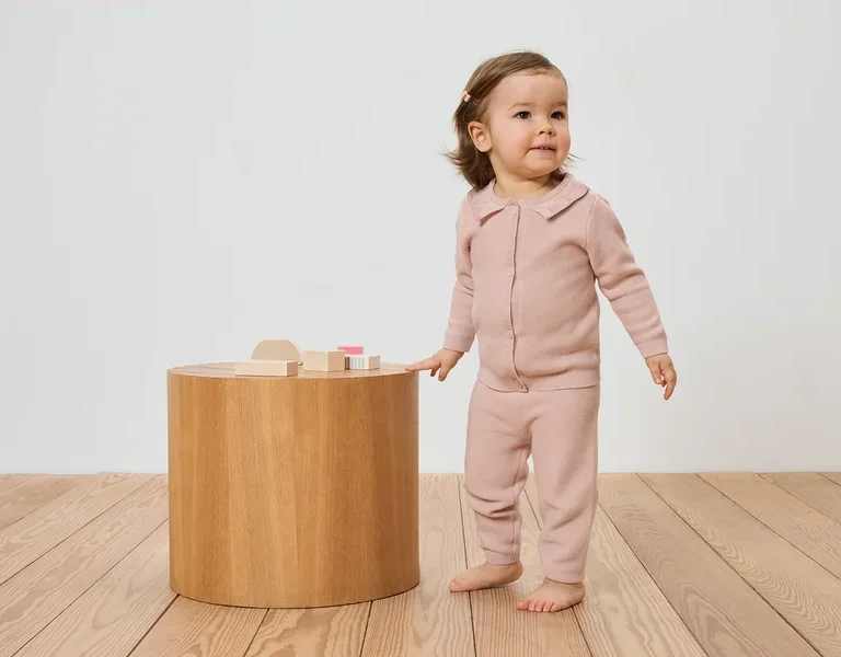 Kleinkind in rosa Strickjacke und Hose steht neben einem Holztisch mit Holzspielzeug auf einem Holzboden.