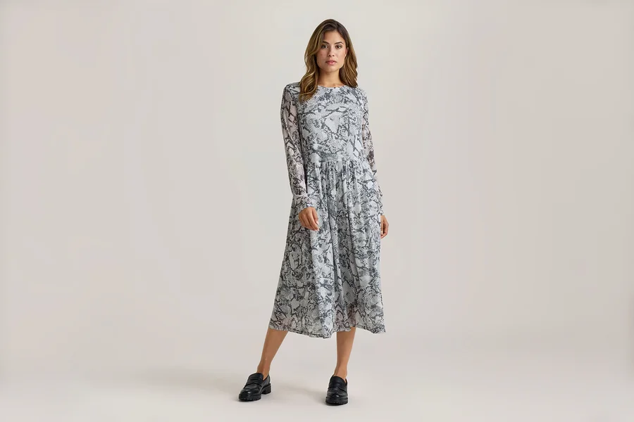 Frau steht in einem langärmeligen Midikleid mit grau-schwarzem Schlangenmuster und schwarzen Loafern.