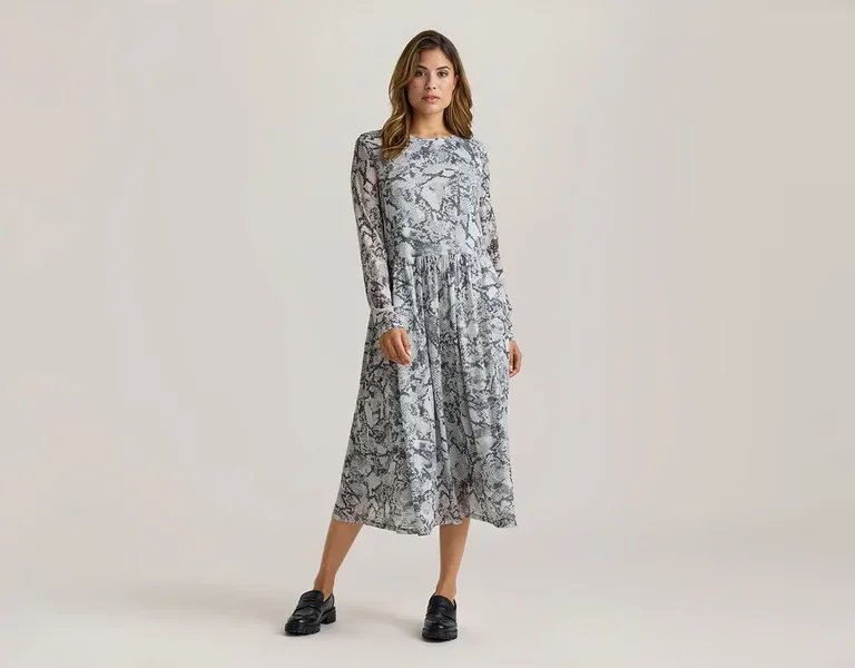 Frau steht in einem langärmeligen Midikleid mit grau-schwarzem Schlangenmuster und schwarzen Loafern.