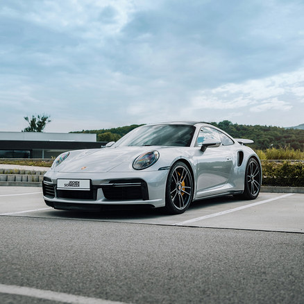 Silberner Porsche mit "Jochen Schweizer" Kennzeichen auf einem Parkplatz, vor einem Gebäude und grünen Hügeln unter bewölktem Himmel.