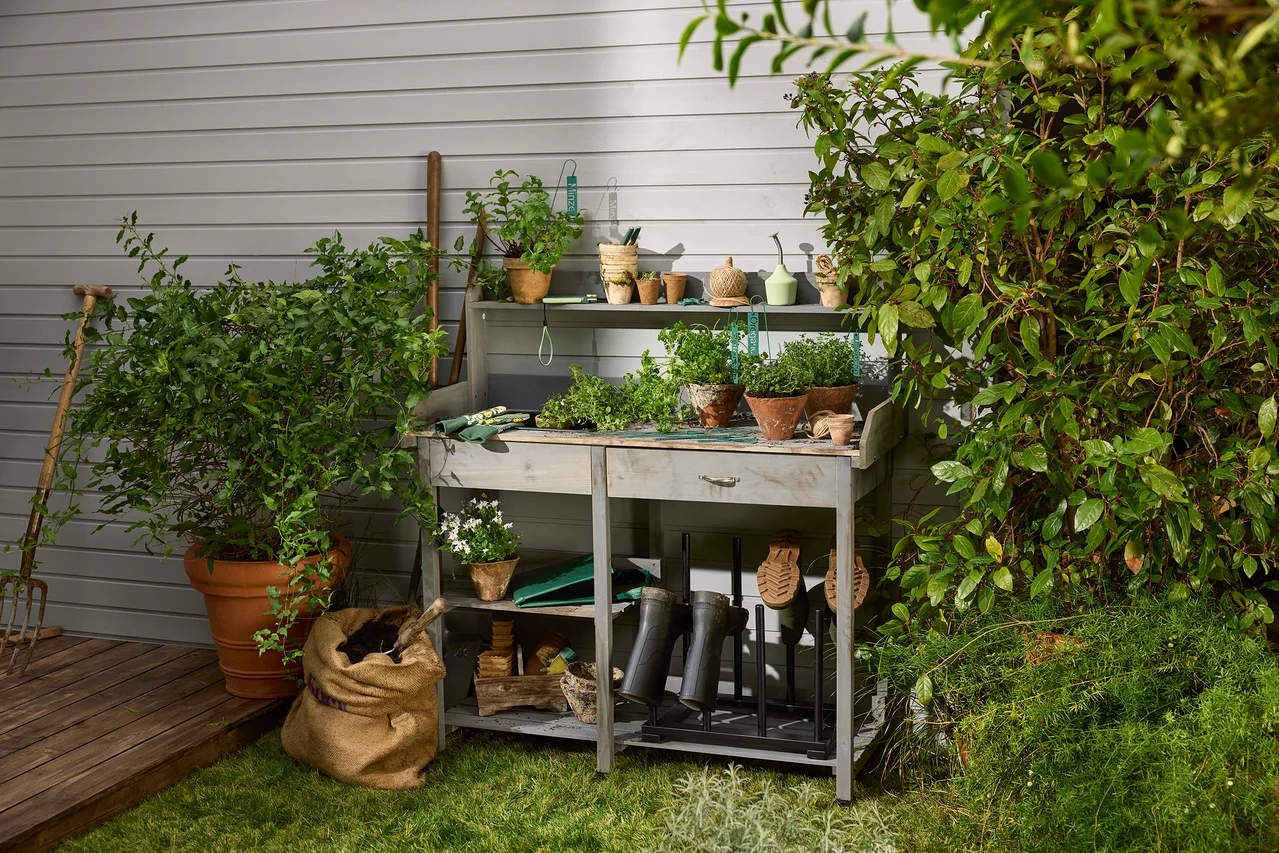 Auf einem Tischchen stehen Blumentöpfe und Gartenzubehör. Im unteren Regal befindet sich ein Stiefelhalter und Kräuterschilder. Eine Anzucht-Gartenbrause ist auch sichtbar.