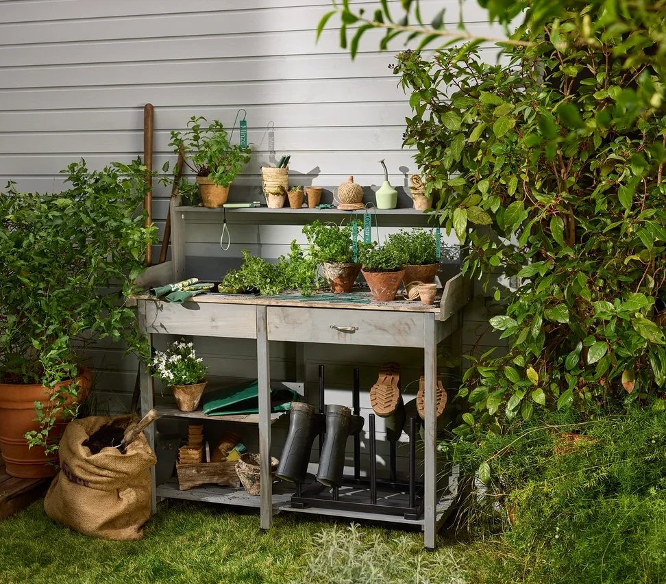 Auf einem Tischchen stehen Blumentöpfe und Gartenzubehör. Im unteren Regal befindet sich ein Stiefelhalter und Kräuterschilder. Eine Anzucht-Gartenbrause ist auch sichtbar.