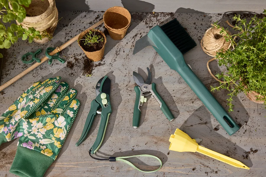 Gartenhandschuhe, 2 Mini-Pflegescheren, 2 Gartenscheren, eine Garten-Fugen-Reinigungsbürste und 3 automatische Bewässerungssticks liegen auf einem Tisch.