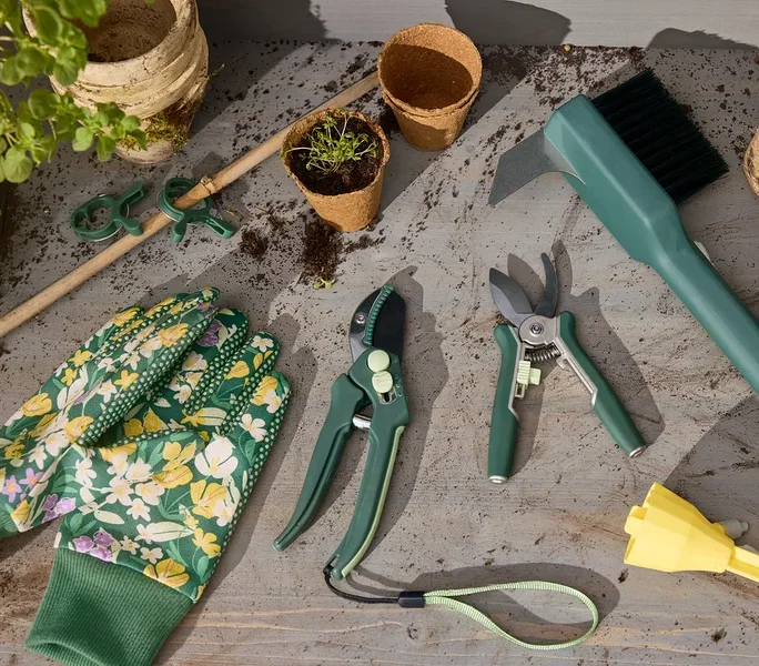 Gartenhandschuhe, 2 Mini-Pflegescheren, 2 Gartenscheren, eine Garten-Fugen-Reinigungsbürste und 3 automatische Bewässerungssticks liegen auf einem Tisch.