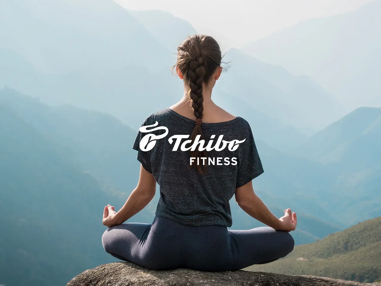 Frau mit Zopf sitzt in Meditationshaltung auf einem Felsen mit Blick auf Berge. Sie trägt ein dunkles T-Shirt mit „Tchibo FITNESS“-Logo und dunkle Leggings.