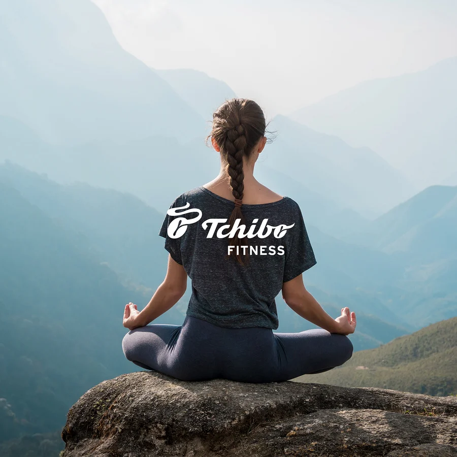 Frau mit Zopf sitzt in Meditationshaltung auf einem Felsen mit Blick auf Berge. Sie trägt ein dunkles T-Shirt mit „Tchibo FITNESS“-Logo und dunkle Leggings.