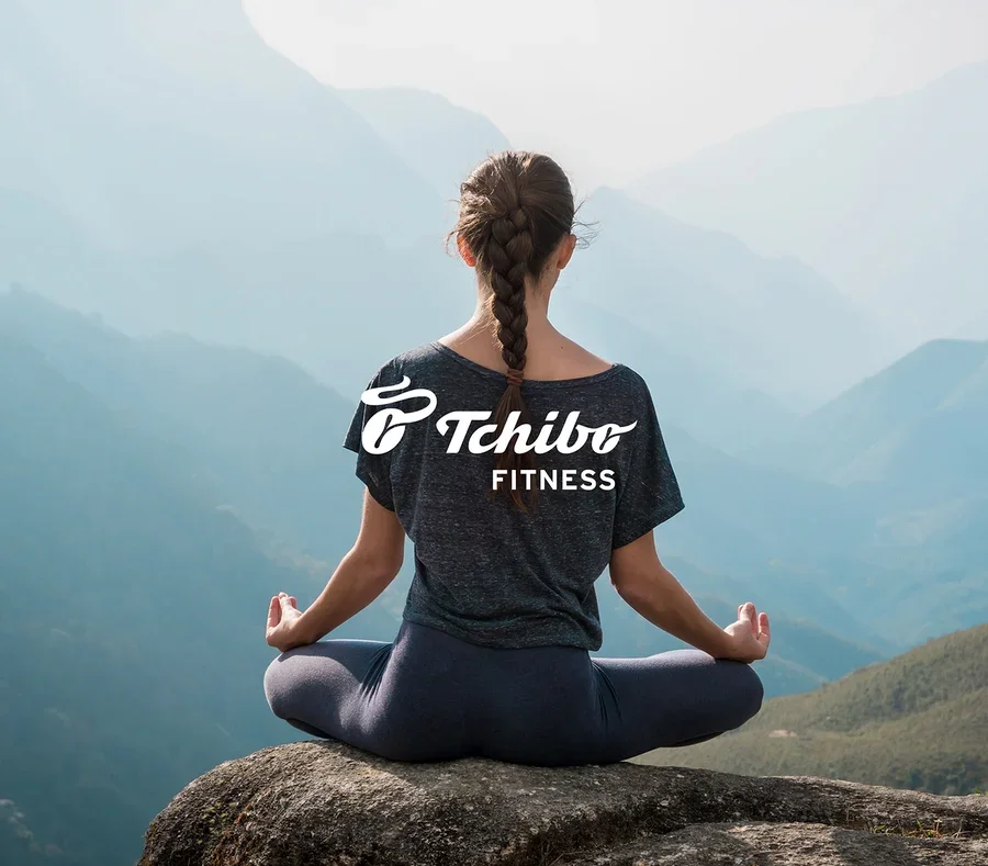 Frau mit Zopf sitzt in Meditationshaltung auf einem Felsen mit Blick auf Berge. Sie trägt ein dunkles T-Shirt mit „Tchibo FITNESS“-Logo und dunkle Leggings.