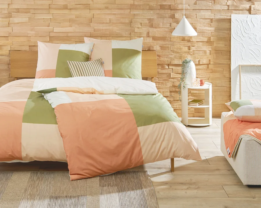 Schlafzimmer mit einem Bett mit karierter Bettwäsche in den Farben Pfirsich, Grün und Creme. Auf einem weißen Nachttisch stehen eine Vase und Dekoration.