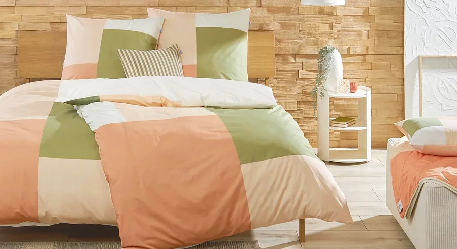 Schlafzimmer mit einem Bett mit karierter Bettwäsche in den Farben Pfirsich, Grün und Creme. Auf einem weißen Nachttisch stehen eine Vase und Dekoration.