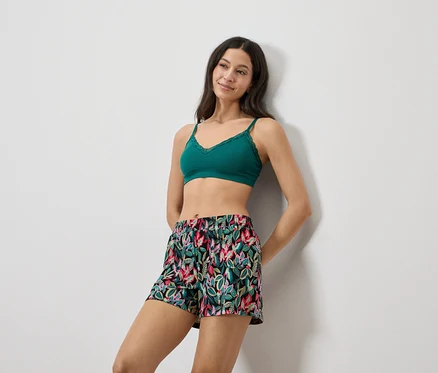 Frau lehnt an einer Wand, trägt ein grünes Spitzen-Bralette und Shorts mit Blumendruck.