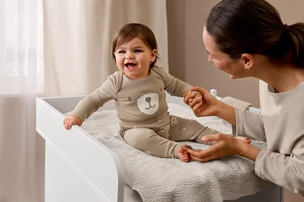Lachendes Baby in beigem, meliertem Feinstrickpullover mit Bärenmotiv und passender Hose sitzt auf Wickelkommode »Leira«, hält die Hand eines Erwachsenen.