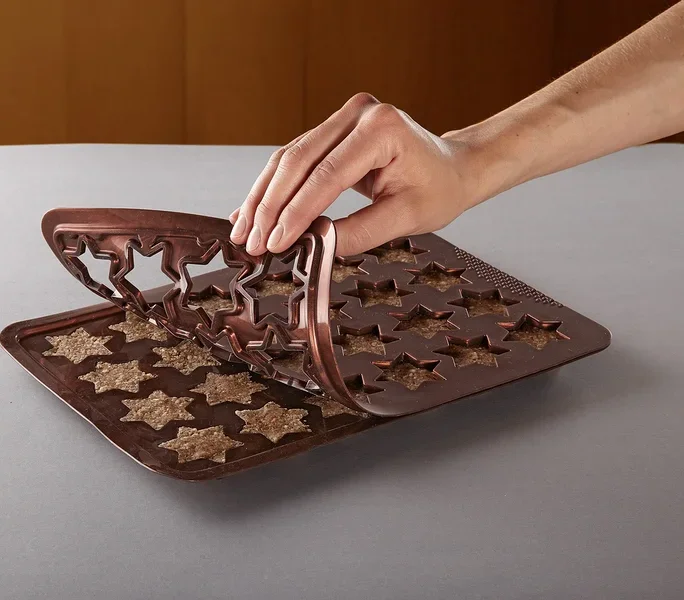 Eine Hand entfernt eine braune Silikon-Backform in Sternform von einem Blech mit sternförmigen Keksen.
