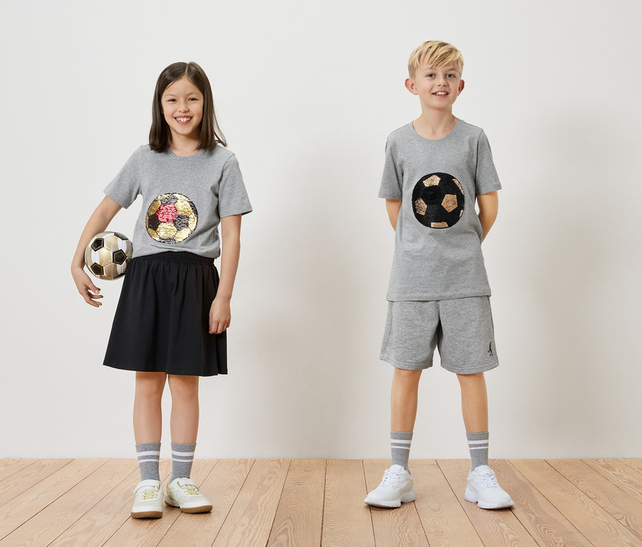Mädchen hält einen Fußball neben einem Jungen, beide tragen graue T-Shirts mit Fußballmotiven.