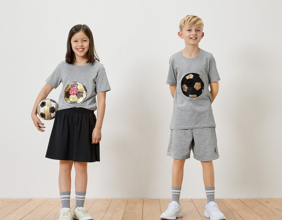 Mädchen hält einen Fußball neben einem Jungen, beide tragen graue T-Shirts mit Fußballmotiven.