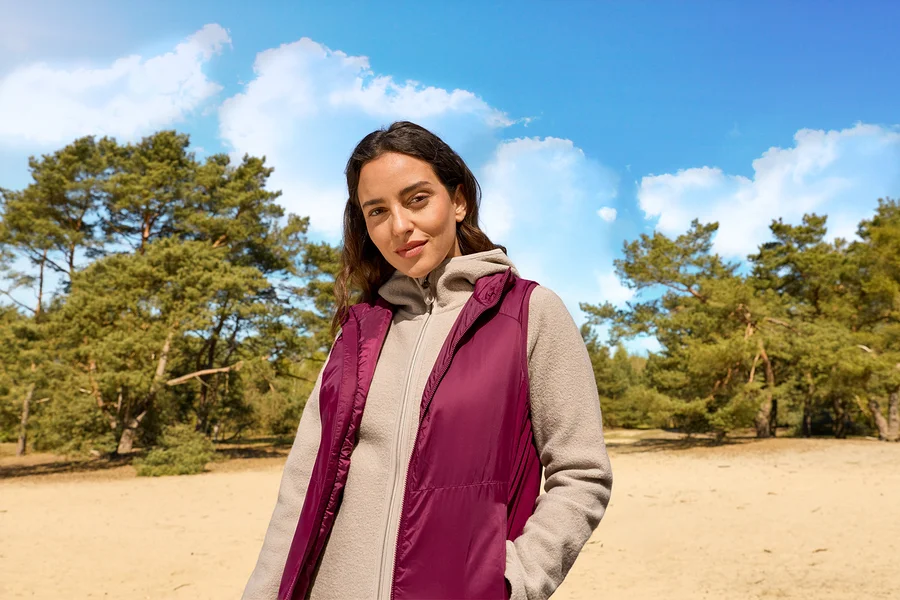 Frau in bordeauxroter Weste und hellbeiger Kapuzen-Fleecejacke steht in sandiger Waldlandschaft unter blauem Himmel.