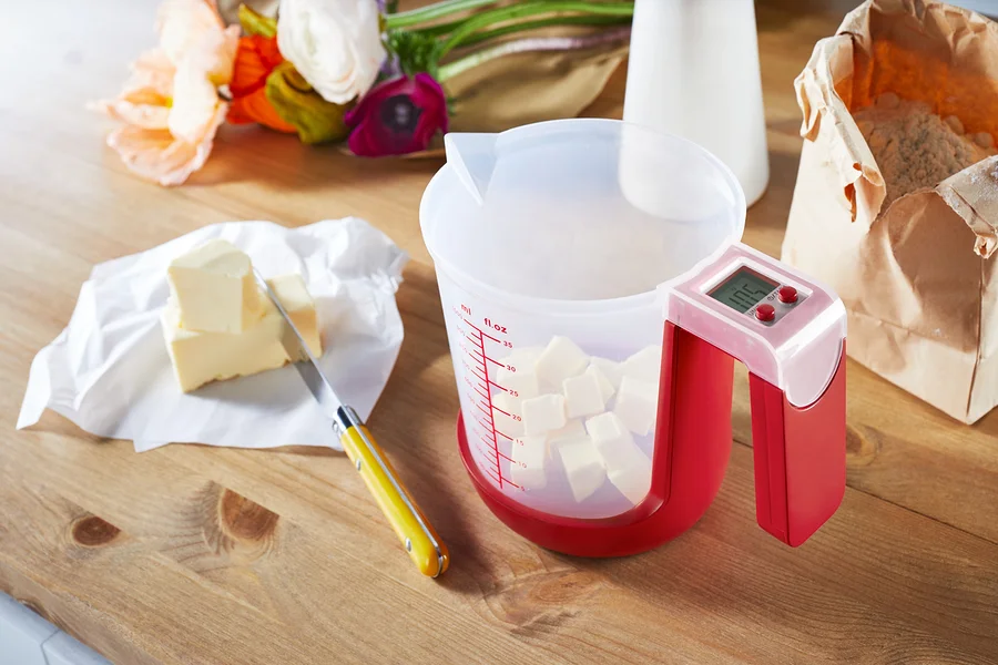 Auf einem Holztisch stehen eine Messbecher-Waage mit gewürfelte Butter, Butter auf Papier und ein Messer.