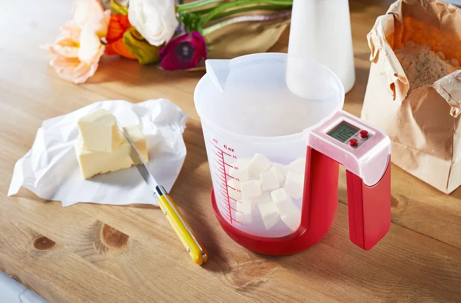 Auf einem Holztisch stehen eine Messbecher-Waage mit gewürfelte Butter, Butter auf Papier und ein Messer.