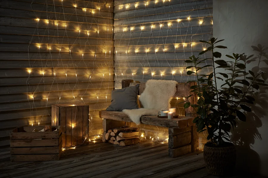 Eine Holzbank mit Kissen und Fell steht vor einer Wand, die mit einem LED-Outdoor-Lichtnetz geschmückt ist.