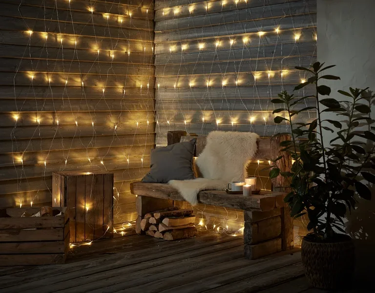 Eine Holzbank mit Kissen und Fell steht vor einer Wand, die mit einem LED-Outdoor-Lichtnetz geschmückt ist.
