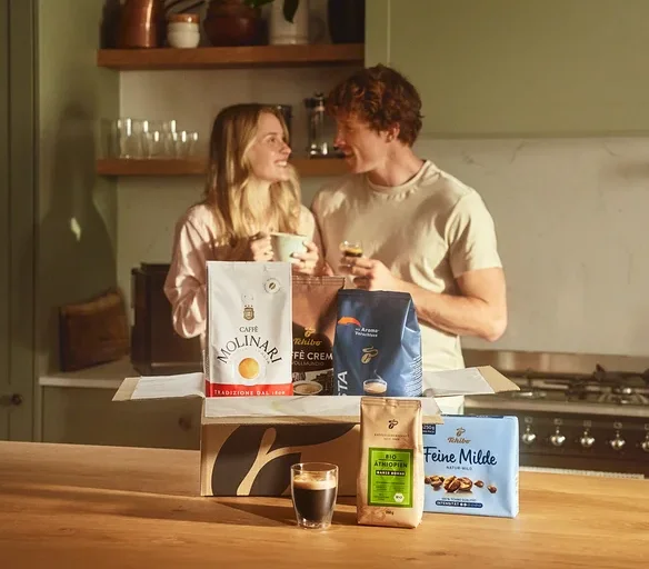 Zwei Personen stehen in einer Küche und halten Kaffeetassen. Vor ihnen steht eine offene Kiste mit verschiedenen Kaffeesorten, darunter Molinari und Tchibo.