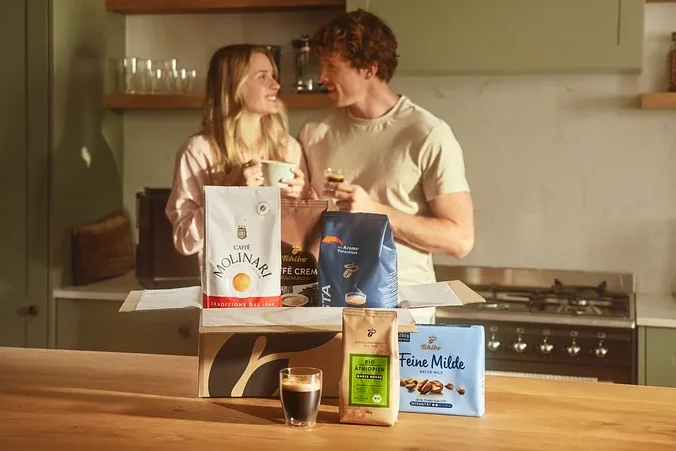 Zwei Personen stehen in einer Küche und halten Kaffeetassen. Vor ihnen steht eine offene Kiste mit verschiedenen Kaffeesorten, darunter Molinari und Tchibo.