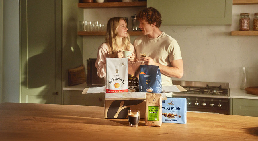 Zwei Personen stehen in einer Küche und halten Kaffeetassen. Vor ihnen steht eine offene Kiste mit verschiedenen Kaffeesorten, darunter Molinari und Tchibo.