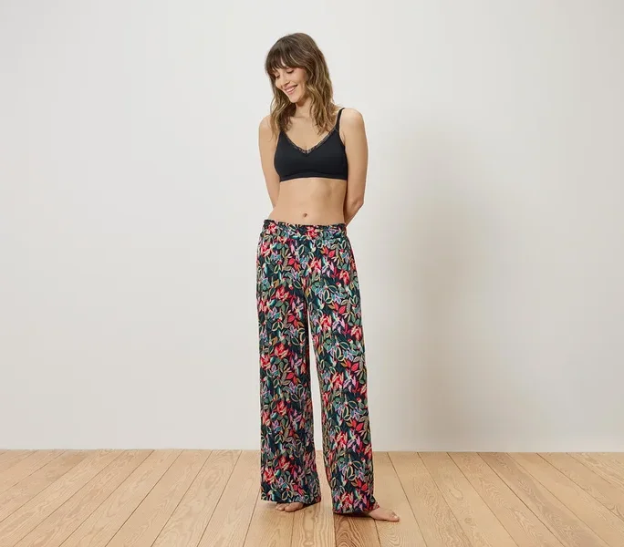 Frau mit schwarzem Bralette und geblümter Hose steht vor einer weißen Wand.