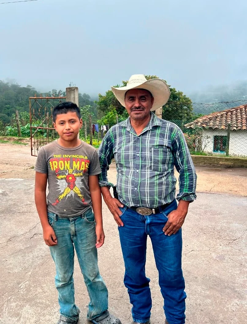 Ein erwachsener Mann mit Cowboyhut und ein Junge in einem Iron Man T-Shirt stehen nebeneinander im Freien. Der Mann trägt ein kariertes Hemd und Jeans, der Junge hellblaue Jeans. Sie stehen auf einem gepflasterten Untergrund vor einer ländlichen Landschaft mit Hügeln und einem Haus.
