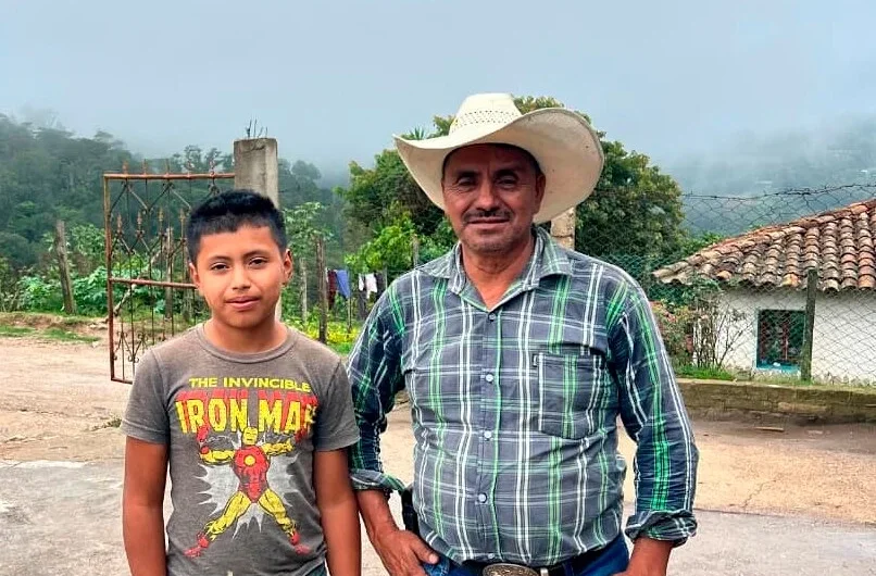 Ein erwachsener Mann mit Cowboyhut und ein Junge in einem Iron Man T-Shirt stehen nebeneinander im Freien. Der Mann trägt ein kariertes Hemd und Jeans, der Junge hellblaue Jeans. Sie stehen auf einem gepflasterten Untergrund vor einer ländlichen Landschaft mit Hügeln und einem Haus.