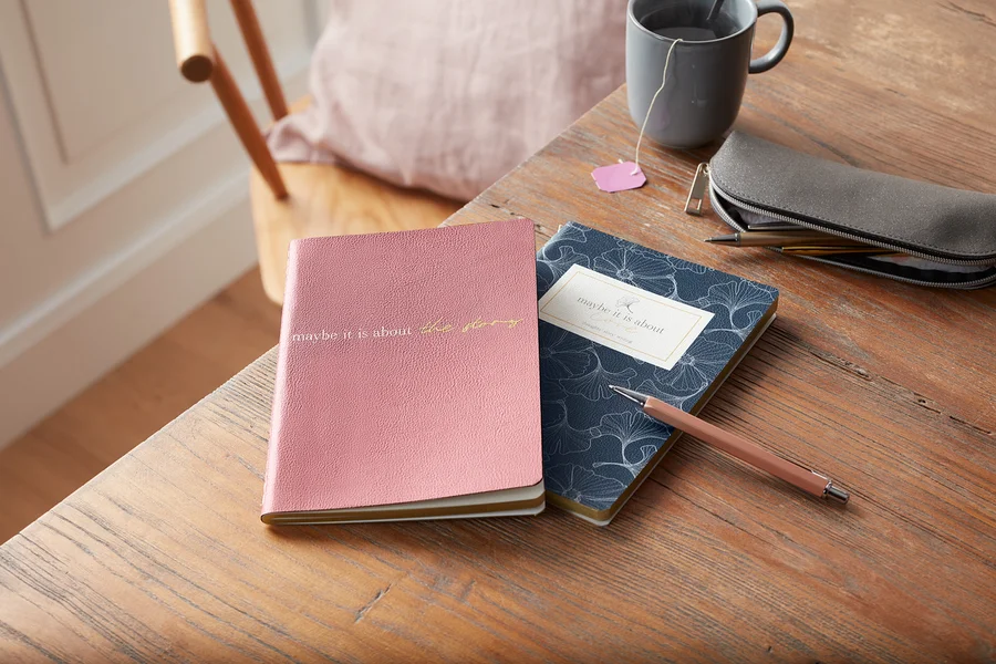 Auf einem Holztisch liegen ein Notizbuch-Set, eine graue Tasse Tee, ein Stifteetui und ein Stift.