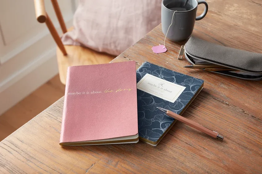 Auf einem Holztisch liegen ein Notizbuch-Set, eine graue Tasse Tee, ein Stifteetui und ein Stift.