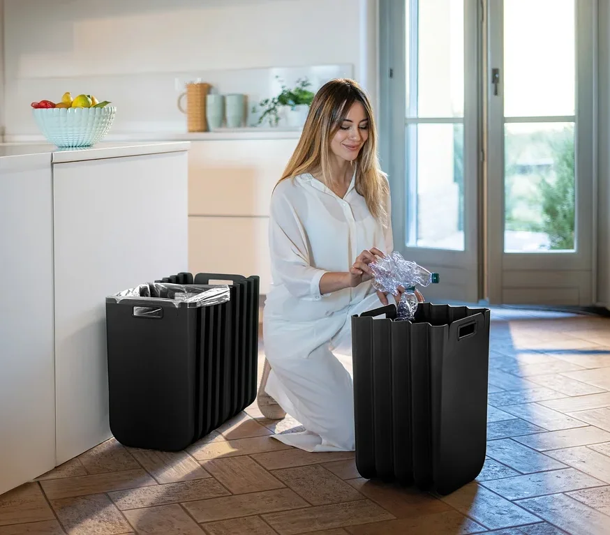 Frau kniet und wirft Plastikflasche in einen von zwei schwarzen Mülleimern im Zimmer.
