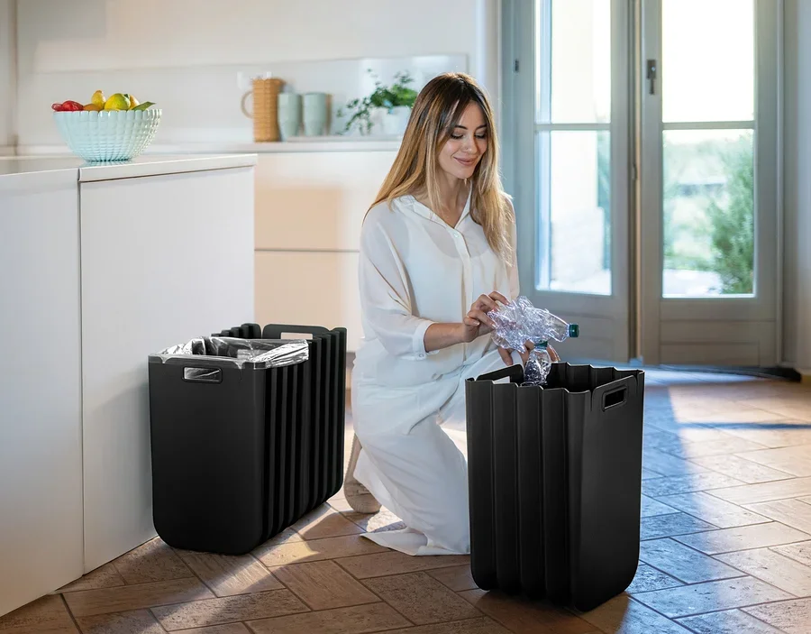 Frau kniet und wirft Plastikflasche in einen von zwei schwarzen Mülleimern im Zimmer.