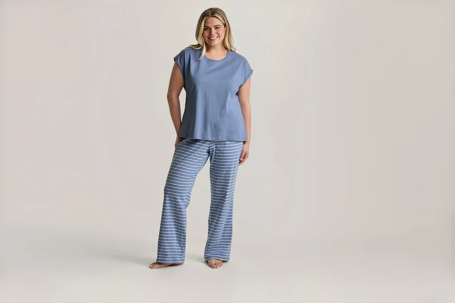 Lächelnde blonde Frau steht barfuß in einem Pyjama-Set mit blauem T-Shirt und blau-weiß gestreiften Hosen.