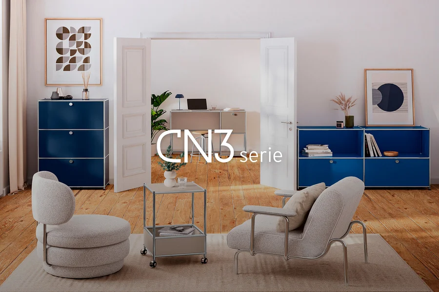 Wohnzimmer mit Holzboden und hellem Teppich. Links ein hoher blauer Modulschrank mit drei Schubladen, rechts ein niedriger blauer Modulschrank mit zwei offenen Fächern und zwei Schubladen. Ein hellgrauer Sessel und ein passender Hocker stehen auf dem Teppich, daneben ein rollbarer Beistelltisch. Im Hintergrund führt eine offene Tür zu einem Büro mit Schreibtisch und Laptop.
