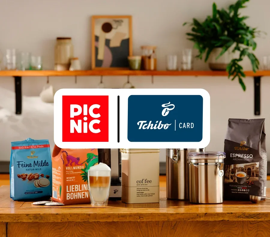 Verschiedene Tchibo Kaffeeprodukte, darunter Pads, ganze Bohnen und ein Latte Macchiato, stehen zusammen mit Kaffeedosen auf einer Holzküchenzeile. Darüber liegen die roten Picnic- und blauen Tchibo Card-Logos.