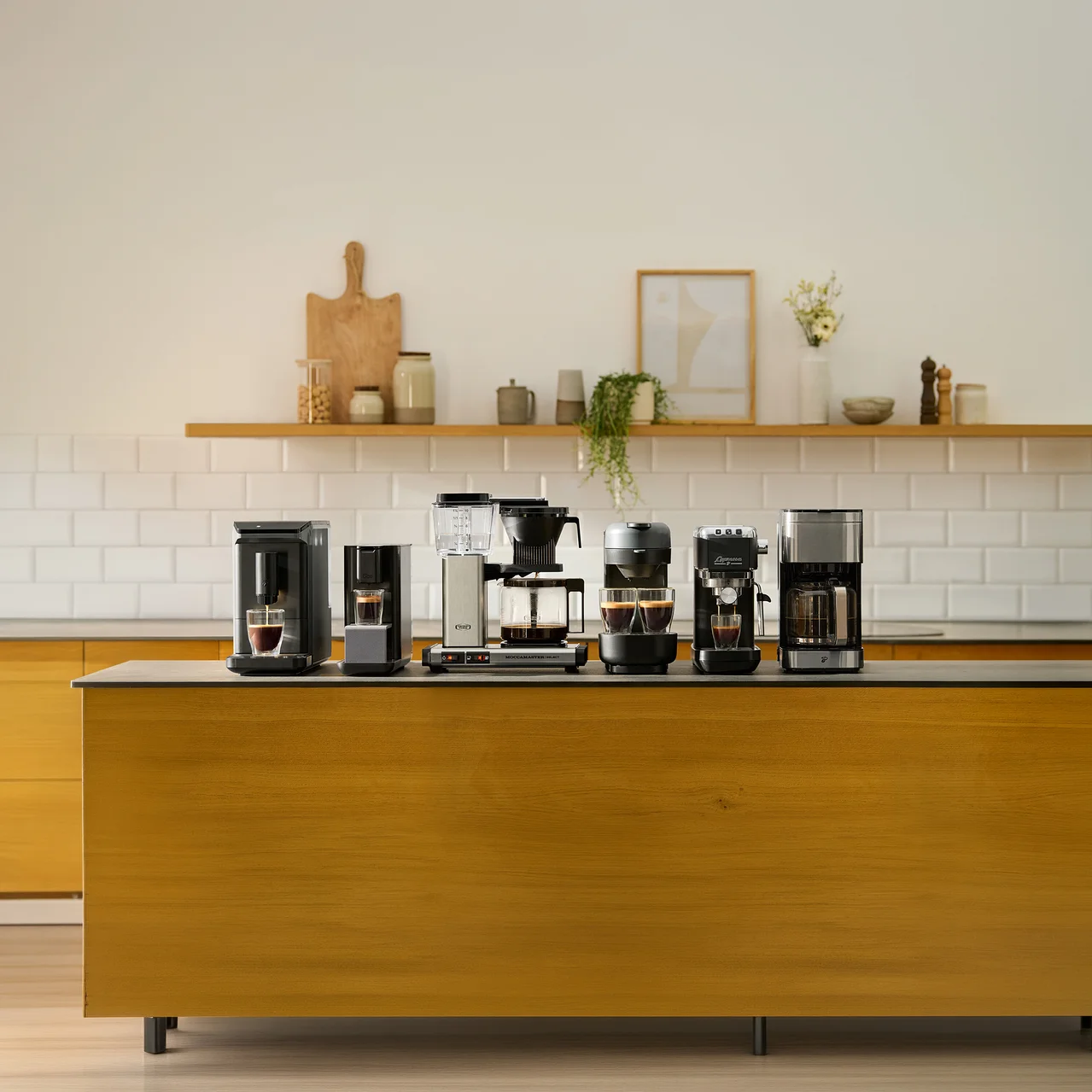 Sechs verschiedene Kaffeemaschinen stehen auf einer dunklen Kücheninsel-Arbeitsplatte. Von links nach rechts sind zwei schwarze Einzelportionsmaschinen, eine silber-schwarze Filterkaffeemaschine, eine dunkelgraue Maschine mit zwei Tassen, eine schwarze Espressomaschine und eine weitere silber-schwarze Filterkaffeemaschine zu sehen. Einige Maschinen haben Tassen mit Kaffee darunter. Im Hintergrund befindet sich ein Holzregal mit Küchendekoration.