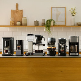 Sechs verschiedene Kaffeemaschinen stehen auf einer dunklen Kücheninsel-Arbeitsplatte. Von links nach rechts sind zwei schwarze Einzelportionsmaschinen, eine silber-schwarze Filterkaffeemaschine, eine dunkelgraue Maschine mit zwei Tassen, eine schwarze Espressomaschine und eine weitere silber-schwarze Filterkaffeemaschine zu sehen. Einige Maschinen haben Tassen mit Kaffee darunter. Im Hintergrund befindet sich ein Holzregal mit Küchendekoration.