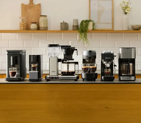 Sechs verschiedene Kaffeemaschinen stehen auf einer dunklen Kücheninsel-Arbeitsplatte. Von links nach rechts sind zwei schwarze Einzelportionsmaschinen, eine silber-schwarze Filterkaffeemaschine, eine dunkelgraue Maschine mit zwei Tassen, eine schwarze Espressomaschine und eine weitere silber-schwarze Filterkaffeemaschine zu sehen. Einige Maschinen haben Tassen mit Kaffee darunter. Im Hintergrund befindet sich ein Holzregal mit Küchendekoration.