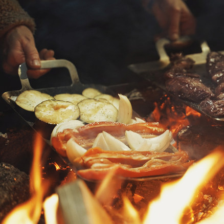 Zwei Hände halten Grillpfannen über offenem Feuer, darauf brutzeln Kartoffelscheiben, Zwiebeln, Paprika und Fleisch.