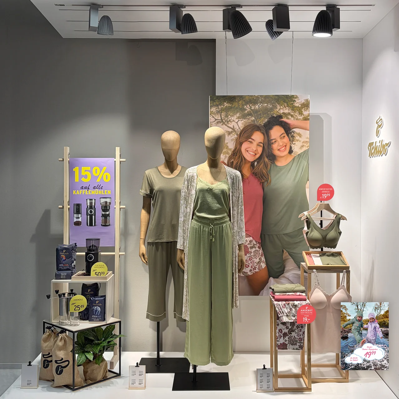 Schaufensterauslage mit zwei Schaufensterpuppen in Loungewear und einem Regal mit Kaffeemühlen und Kaffeebohnen. Links ein Schild „15% auf alle KAFFEEMÜHLEN“. Die linke Puppe trägt ein olivgrünes Pyjama-Set, die rechte eine hellgrüne Camisole, weite Hose und einen floralen Cardigan. Im Hintergrund ein Poster mit zwei lächelnden Frauen. Rechts ein Ständer mit einem grünen BH, einer rosa Camisole und gefalteter Kleidung.