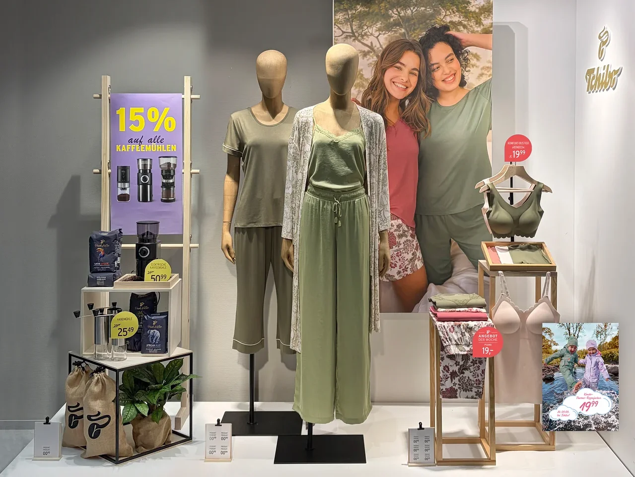 Schaufensterauslage mit zwei Schaufensterpuppen in Loungewear und einem Regal mit Kaffeemühlen und Kaffeebohnen. Links ein Schild „15% auf alle KAFFEEMÜHLEN“. Die linke Puppe trägt ein olivgrünes Pyjama-Set, die rechte eine hellgrüne Camisole, weite Hose und einen floralen Cardigan. Im Hintergrund ein Poster mit zwei lächelnden Frauen. Rechts ein Ständer mit einem grünen BH, einer rosa Camisole und gefalteter Kleidung.