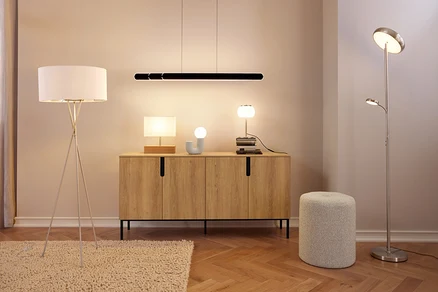 Ein modernes Zimmer mit einem Holz-Sideboard, auf dem drei Lampen und ein Deko-Objekt stehen, unter einer schwarzen linearen Pendelleuchte. Links steht eine weiße Stehlampe mit Stativfuß auf einem Teppich, rechts eine silberne Deckenfluter-Stehlampe mit Leselicht neben einem grauen Hocker.