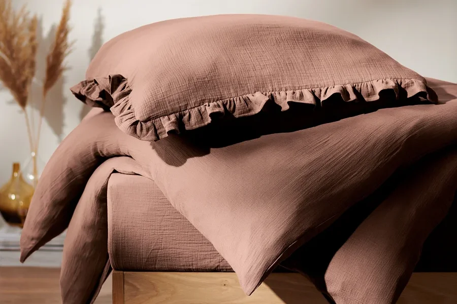 Detailaufnahme von gestapelter Musselin-Bettwäsche auf einem Bett mit Holzrahmen, mit braunen Hausschuhen auf dem Boden.