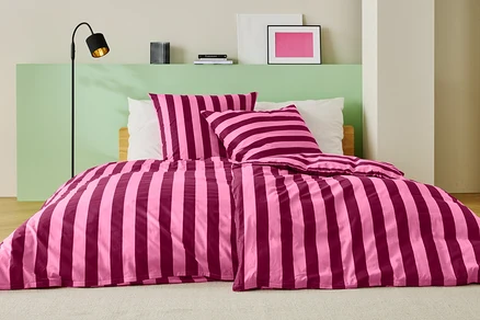 Bett mit rosa-weinroter Streifenbettwäsche, zwei Kissen und einer Stehlampe.