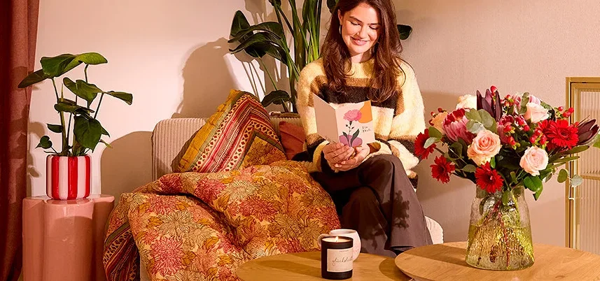 Frau sitzt auf einem Sofa und liest eine Karte. Auf dem Tisch stehen eine Kerze und ein Blumenstrauß.