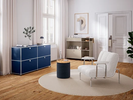 Modernes Wohnzimmer mit Holzboden und weißen Wänden. Links steht ein dunkelblaues Sideboard unter einem Fenster, darauf eine Vase und eine Lampe. In der Mitte liegt ein runder Teppich mit einem zylindrischen Beistelltisch und einer Wasserflasche. Rechts steht ein heller Sessel und ein beiges Rollregal mit Geschirr. An der Wand hängt ein abstraktes Bild.