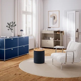 Modernes Wohnzimmer mit Holzboden und weißen Wänden. Links steht ein dunkelblaues Sideboard unter einem Fenster, darauf eine Vase und eine Lampe. In der Mitte liegt ein runder Teppich mit einem zylindrischen Beistelltisch und einer Wasserflasche. Rechts steht ein heller Sessel und ein beiges Rollregal mit Geschirr. An der Wand hängt ein abstraktes Bild.