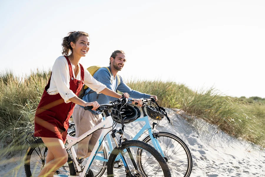 Zwei Personen lächeln und fahren mit Fahrrädern an einem Sandstrand in der Nähe von grasbewachsenen Dünen.