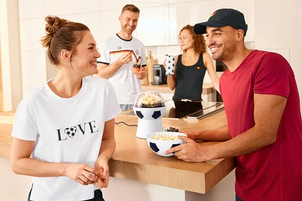 Vier Personen in einer Küche. Ein Mann hält eine mit Popcorn gefüllte Schüssel neben einer Fußball-Popcornmaschine. Eine Frau trägt ein T-Shirt mit Print.
